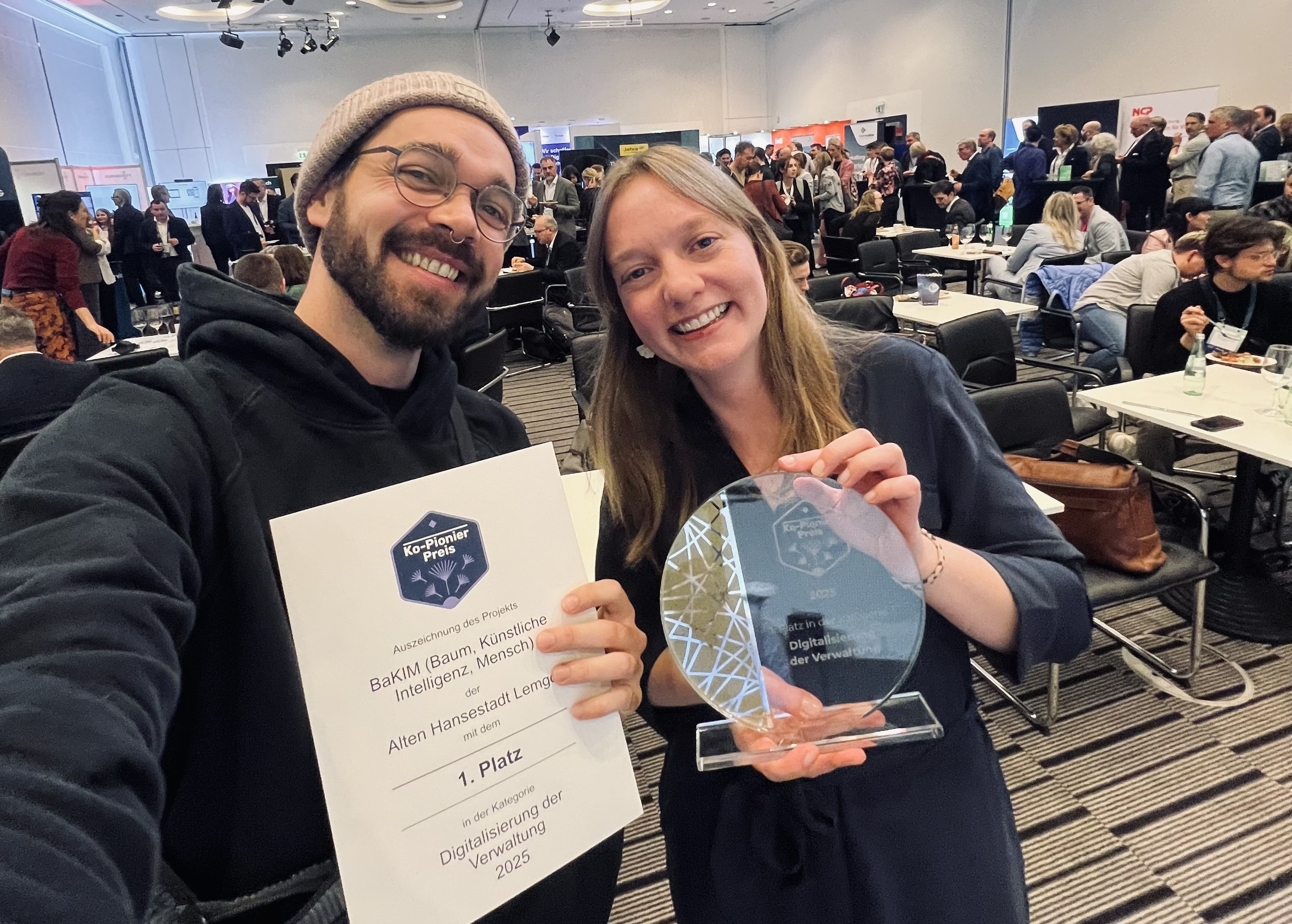 Selfie von einem Mann und einer Frau, die lachend eine Urkunde und einen Preis in die Kamera halten.