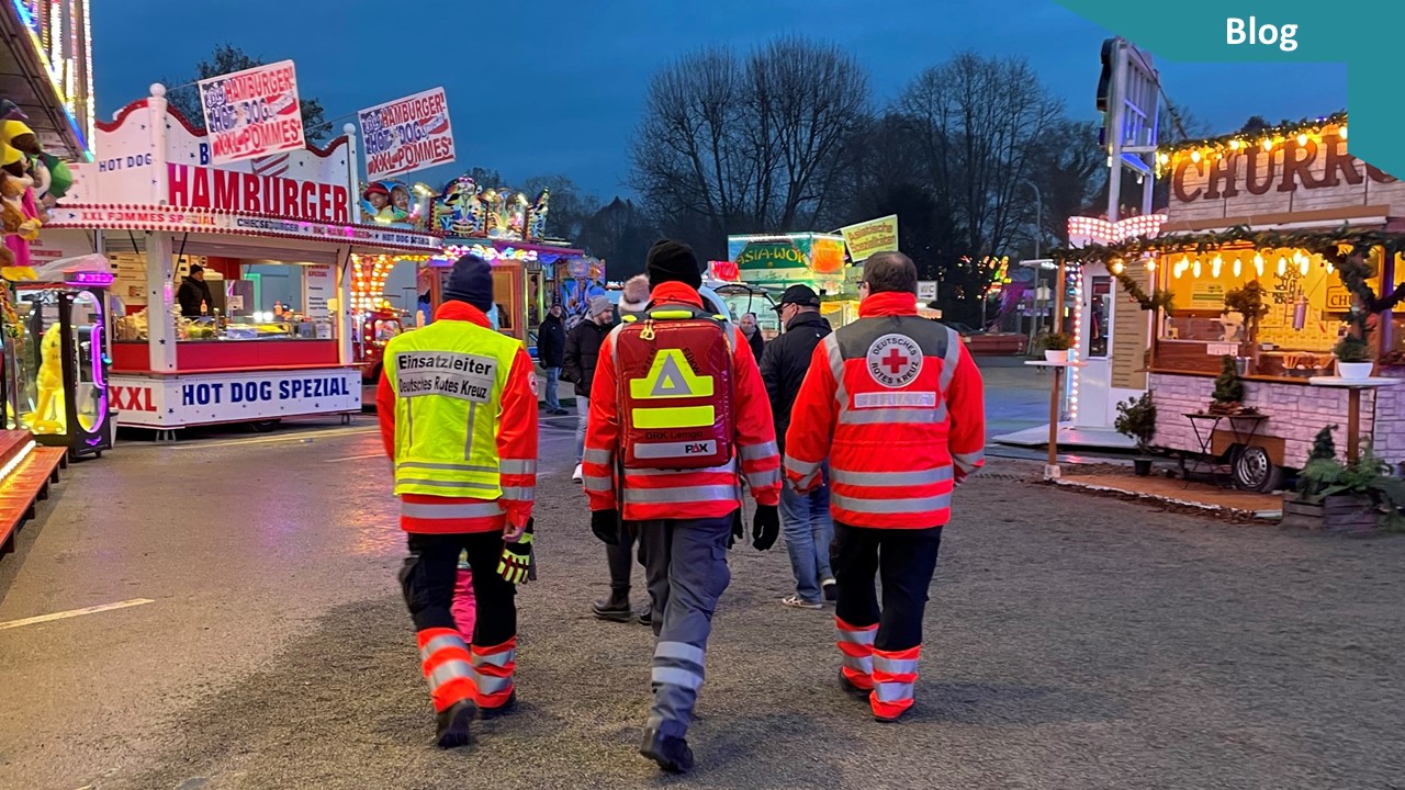 Drei Einsatzkräfte in Uniform des Deutschen Roten Kreuzes von hinten auf einer Kirmes 